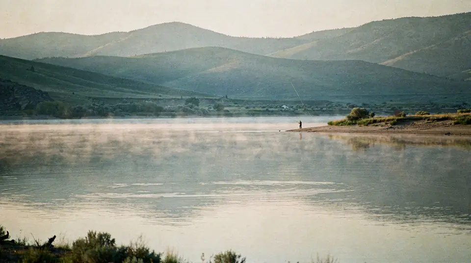 A calm reservoir at sunrise with mist on the water and a distant angler on the shore