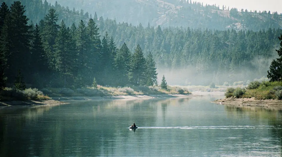 A float tube angler on a calm mountain lake with mist and pines