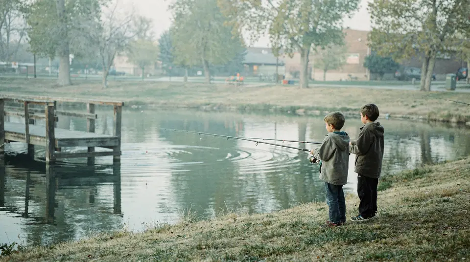 Two children seen from behind fishing from the grassy bank of a small park pond