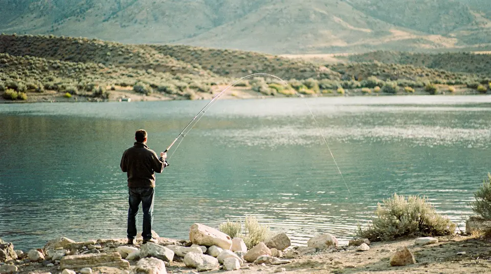 An angler casting from a rocky shore at a wide reservoir with sagebrush hills